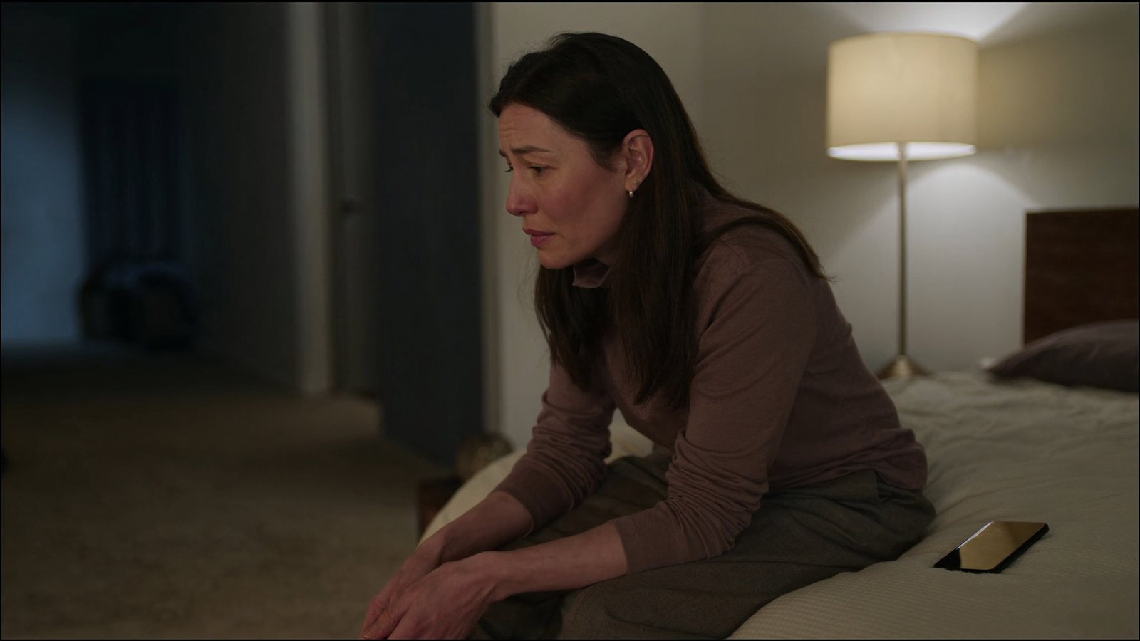 Woman sitting on the edge of her bed in a dimly lit bedroom at night, her phone face down on the duvet beside her, looking away from it after reading her report