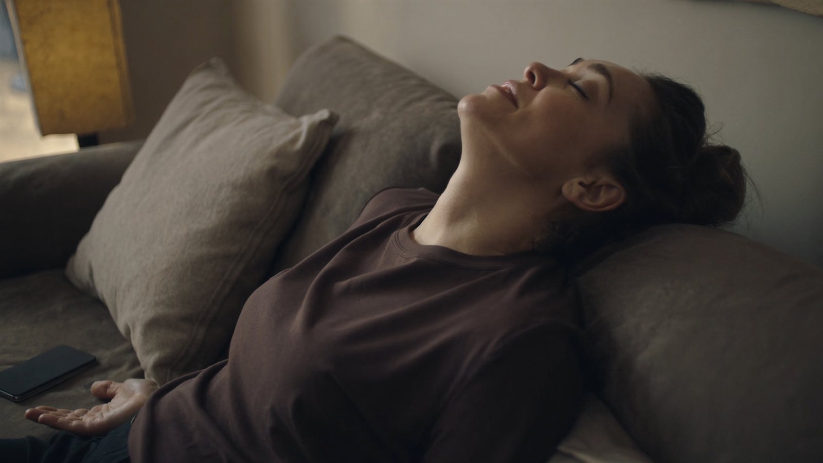 Woman sitting back on a sofa in the evening with her eyes closed, phone set down beside her