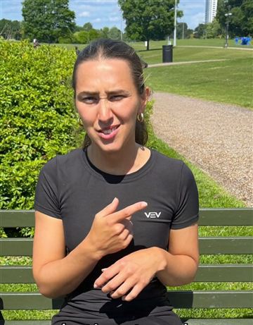 Woman in a black athletic shirt on a park bench pointing at the camera mid sentence