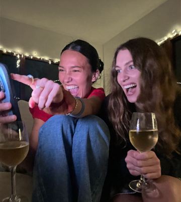 Two friends with wine glasses laughing in disbelief at a phone