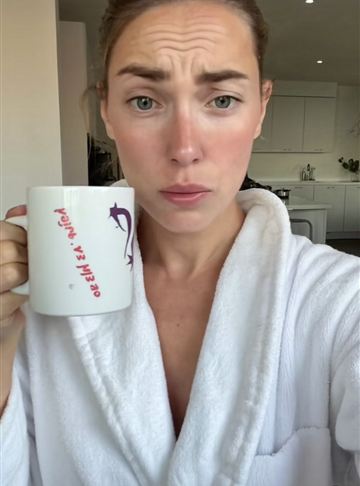 Woman in a white bathrobe holding a coffee mug with an unimpressed look in a bright kitchen