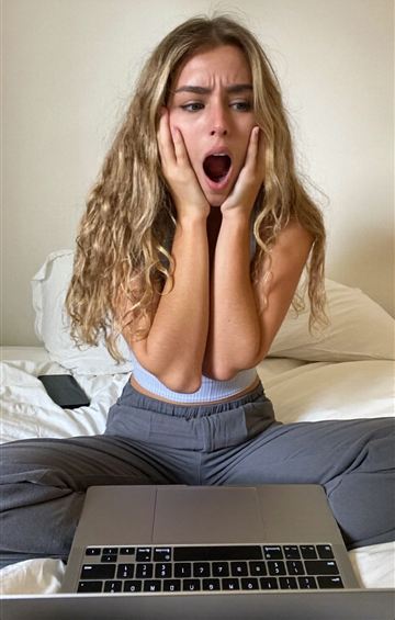 Woman gasping with both hands on her cheeks in front of a laptop on her bed