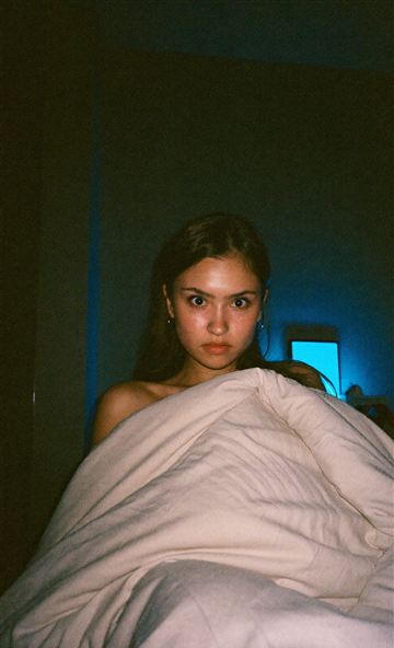 Wide eyed young woman peeking over a duvet in a dark bedroom lit by a blue phone glow